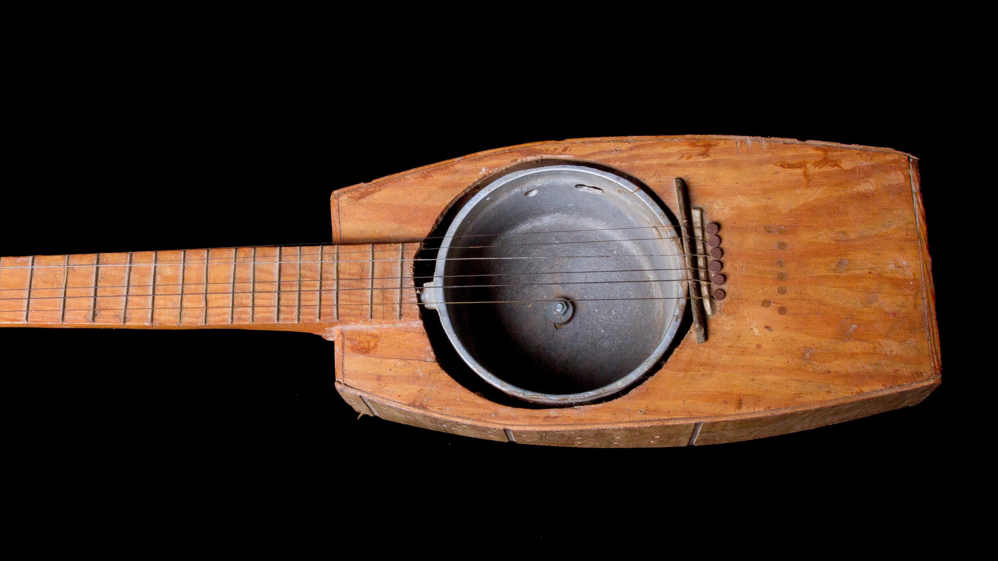 A rustic wooden instrument with 6 guitar strings and an old pot. Rectangularly shaped with irregular frets.