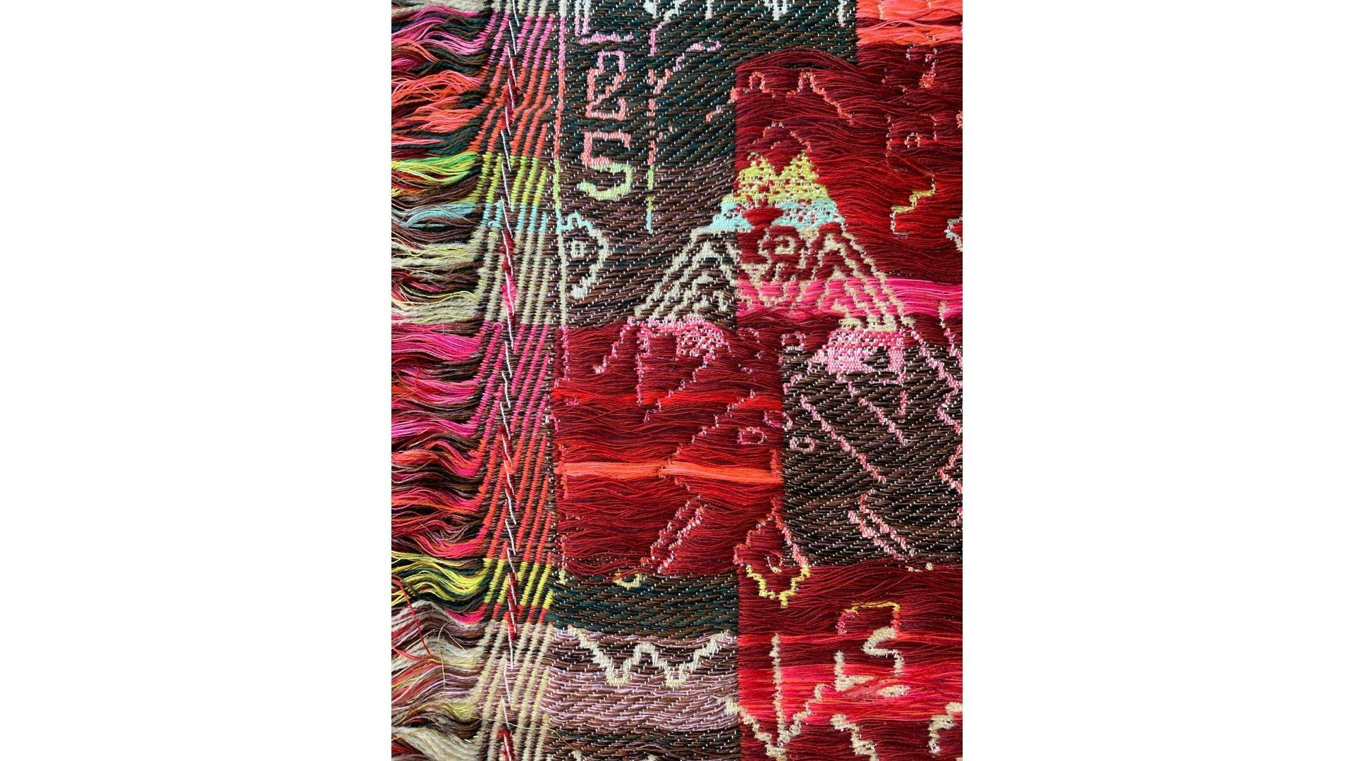 Close-up of a textile in red, gray, green, and orange. The close-up shows a mountain and symbols.